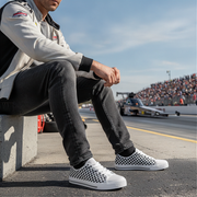 Person sitting on a track wearing checkered shoes with a race track and cars in the background.