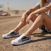 Person wearing checkered shoes sitting on a dirt track with cars in the background