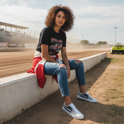 Woman sitting on a concrete barrier at a race track wearing a black t-shirt, jeans, and checkered shoes.
