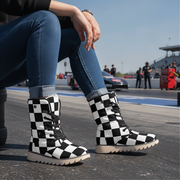 Person wearing checkered boots on a race track with cars and people in the background