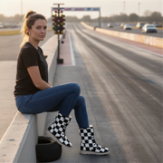Woman sitting on a guardrail by a race track wearing checkered boots.