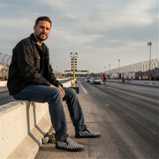 Man sitting on a race track with cars in the background wearing a checkered slip on shoes