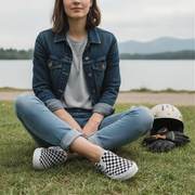 Person sitting on grass wearing denim jacket, jeans, and checkered shoes with a helmet and gloves nearby.