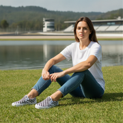 Woman sitting on grass by a lake wearing a white t-shirt and blue jean and white checkered slip on shoes