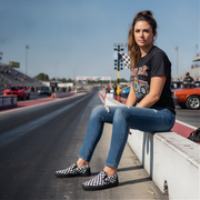 Woman sitting on a race track wearing checkered shoes and a black t-shirt with a graphic design.