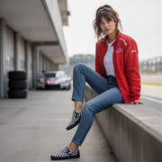 Woman in red jacket and checkered shoes sitting on a concrete ledge with cars in the background