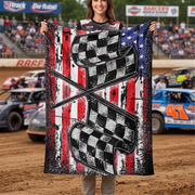 A woman stands on a dirt race track holding up a large printed blanket or throw. The blanket features a distressed American flag design with red, white, and blue vertical stripes, black-and-white checkered flags, and crossed flag motifs.