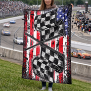 A woman holds up a large printed blanket or tapestry featuring a distressed American flag design with two crossed checkered flags over red, white, and blue stripes. The background shows a racetrack scene with cars and spectators.