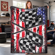 A woman stands in a garage holding up a large printed blanket or tapestry featuring two crossed checkered flags over a distressed American flag background with red, white, and blue stripes and white stars.