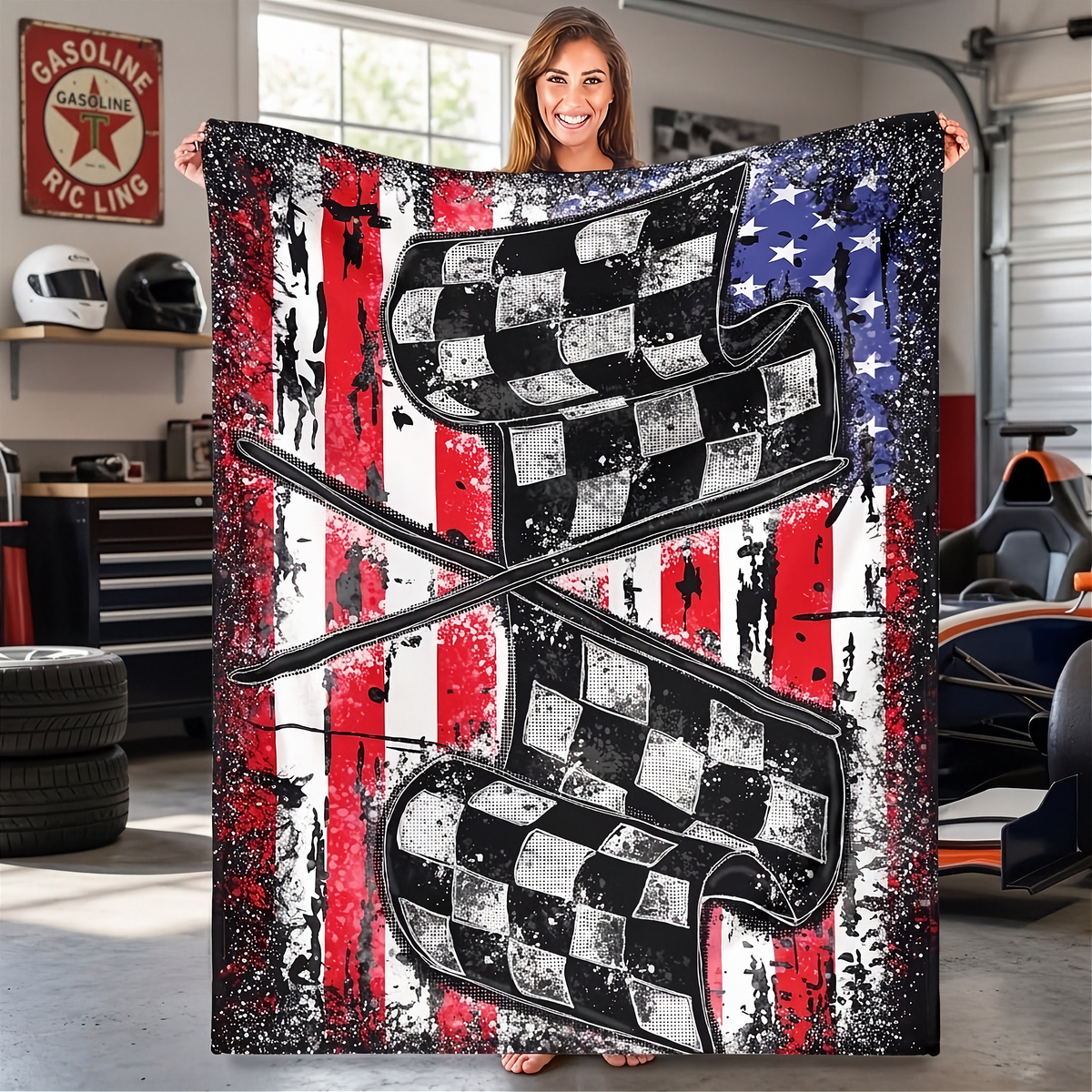 A woman stands in a garage holding up a large printed blanket or tapestry featuring two crossed checkered flags over a distressed American flag background with red, white, and blue stripes and white stars.