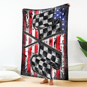 A person holds up a large fleece blanket featuring a distressed American flag design with crossed checkered flags in the center, shown in a bedroom setting.