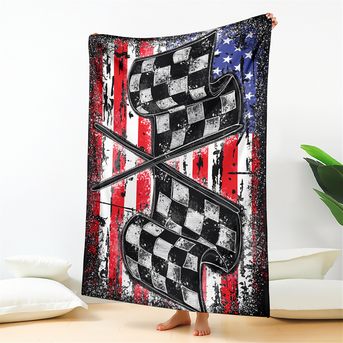 A person holds up a large fleece blanket featuring a distressed American flag design with crossed checkered flags in the center, shown in a bedroom setting.