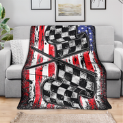 A throw blanket draped over a light gray sofa in a living room setting, featuring a distressed American flag design with red and white stripes, a blue field with white stars, and two large black-and-white checkered race flags crossing in the center.