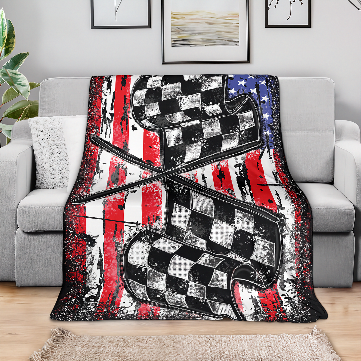 A throw blanket draped over a light gray sofa in a living room setting, featuring a distressed American flag design with red and white stripes, a blue field with white stars, and two large black-and-white checkered race flags crossing in the center.