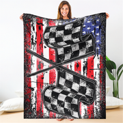 A woman holds up a large blanket or throw featuring a distressed American flag background with two crossed checkered flags in black, white, and gray. The blanket is shown in a bedroom setting with a white wall, pillows, and a plant visible.