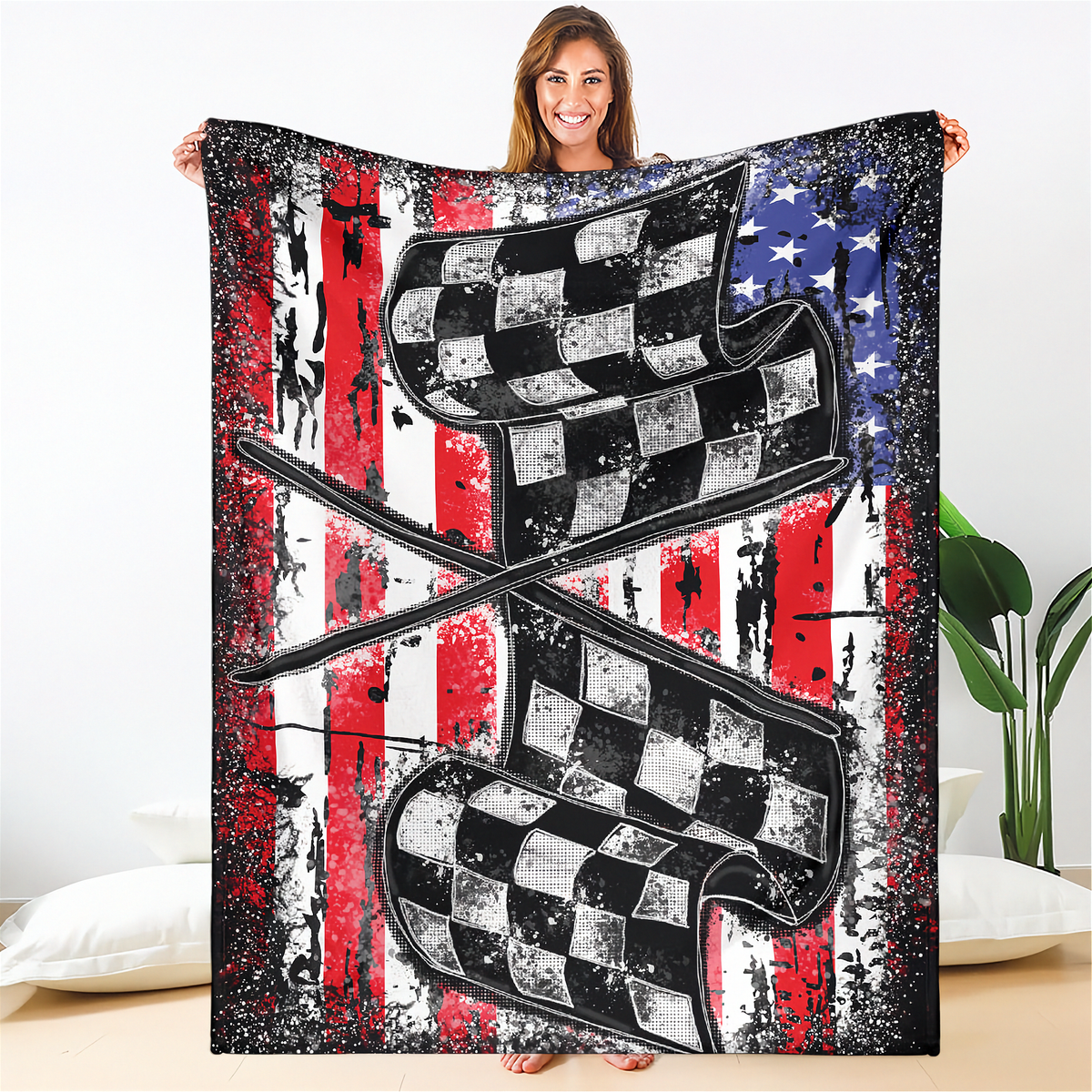 A woman holds up a large blanket or throw featuring a distressed American flag background with two crossed checkered flags in black, white, and gray. The blanket is shown in a bedroom setting with a white wall, pillows, and a plant visible.