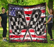 2 women standing on the grass holding up a large square quilt featuring a distressed American flag design with two crossed checkered flags in the center.