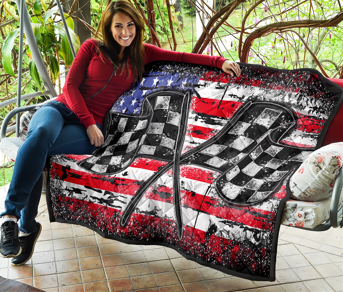 A woman sitting on a porch holding up a large square quilt featuring a distressed American flag design with two crossed checkered flags in the center.
