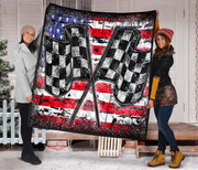 2 women standing on a snowy porch holding up a large square quilt featuring a distressed American flag design with two crossed checkered flags in the center.