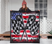 A woman standing on a snowy porch holds up a large square quilt featuring a distressed American flag design with two crossed checkered flags in the center.