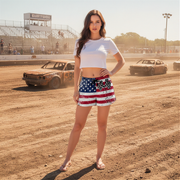 Woman in a white top and American flag skirt standing on a dirt track with vintage cars in the background.