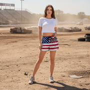 Woman in a white crop top and American flag shorts standing on a dirt track with cars in the background.