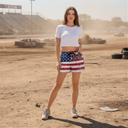 Woman in a white crop top and American flag shorts standing on a dirt track with cars in the background.
