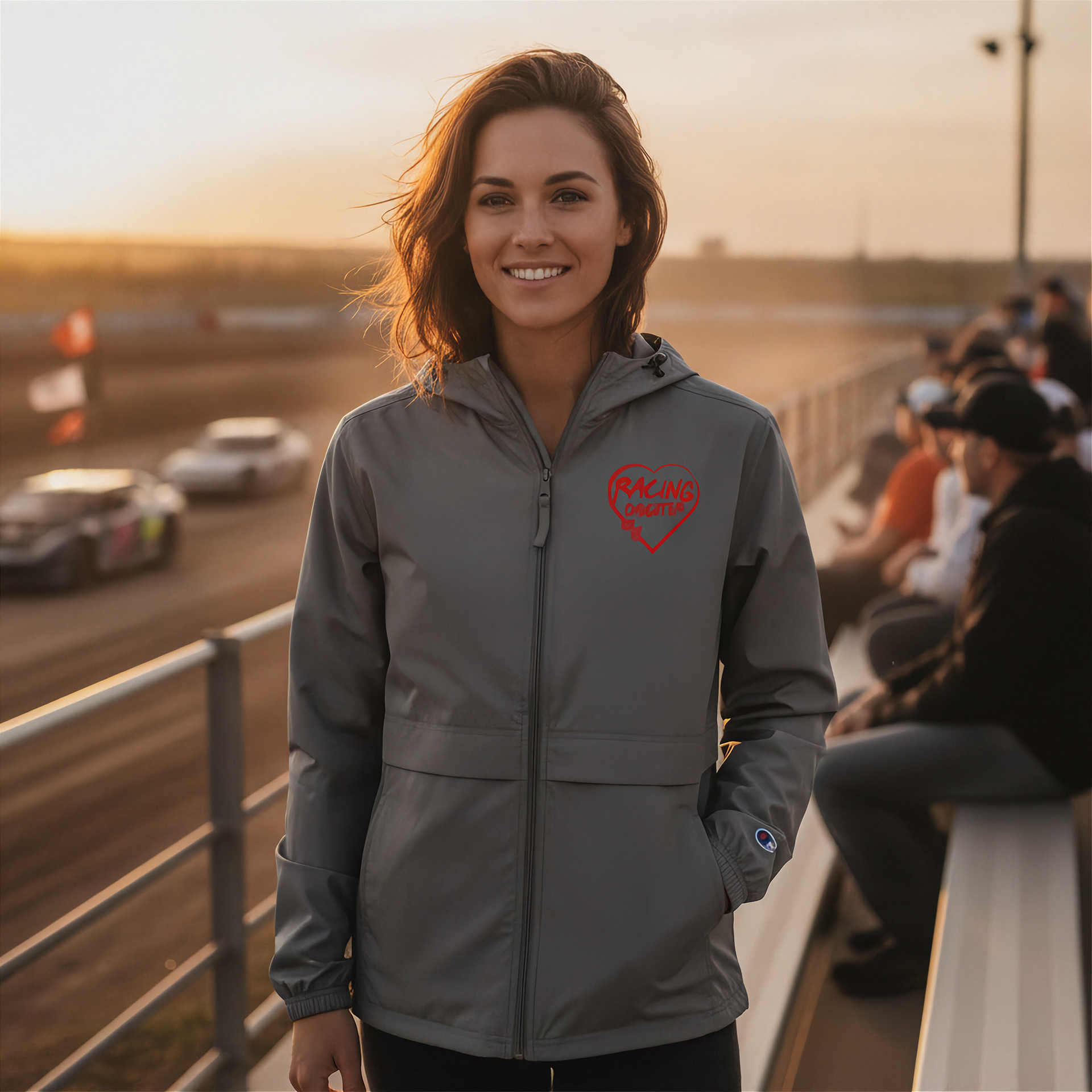 Woman wearing a gray jacket with a red racing daughter heart design at a race track during sunset.