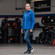 Woman in a blue jacket with a red racing daughter heart design standing in a garage with racers in the background