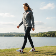 Woman walking by a lake wearing a gray jacket with a red racing daughter heart design and black pants.