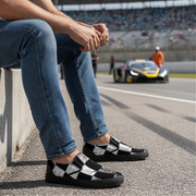 Man sitting on a race track wearing checkered shoes with a car in the background.