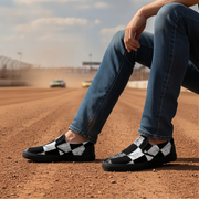 Person wearing black and white checkered shoes on a dirt track with cars in the background.