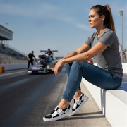 Woman sitting on a track wearing black and white checkered slip on shoes with a race car in the background.