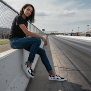 woman sitting on a track barrier at a race track wearing checkered slip on shoes