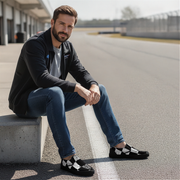 Man sitting on a concrete block by a race track wearing black checkered slip on shoes.