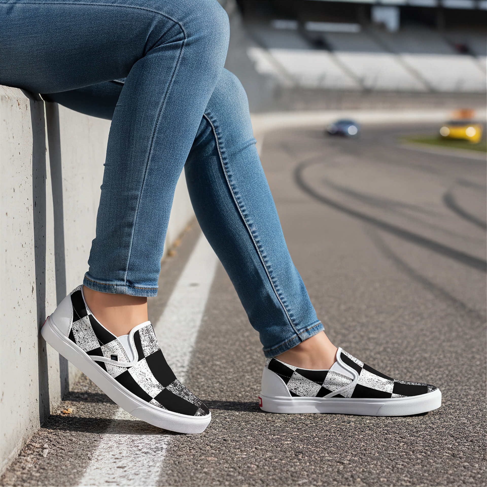 Woman wearing black and white checkered shoes with blue jeans sitting on a curb by a race track.