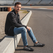 Man sitting on a race track wearing a black jacket, blue jeans, and black checkered shoes.