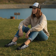 Person sitting on grass by a lake wearing a cap and casual clothing and checkered shoes.