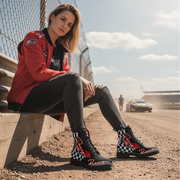 Woman sitting on a race track wearing a red racing jacket and black checkered boots.