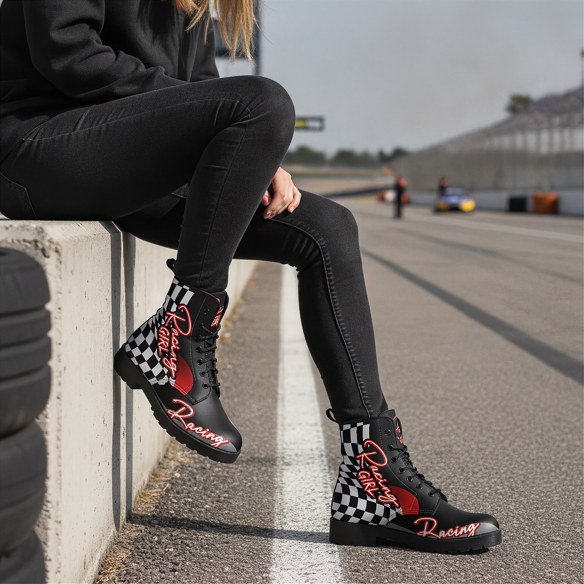 Woman sitting on a race track wearing black racing boots with checkered patterns and red text.