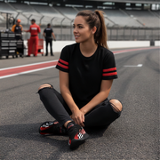 Woman sitting on a race track wearing black t-shirt with red stripes and black shoes with checkered design.