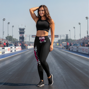 Woman in racing gear standing on a race track with spectators and cars in the background