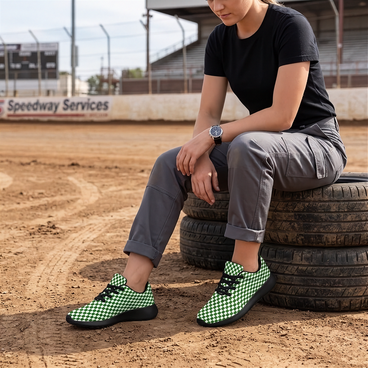 A person sitting on stacked tires at a dirt track wearing green and black checkered sneakers, a black t-shirt, and gray cargo pants.