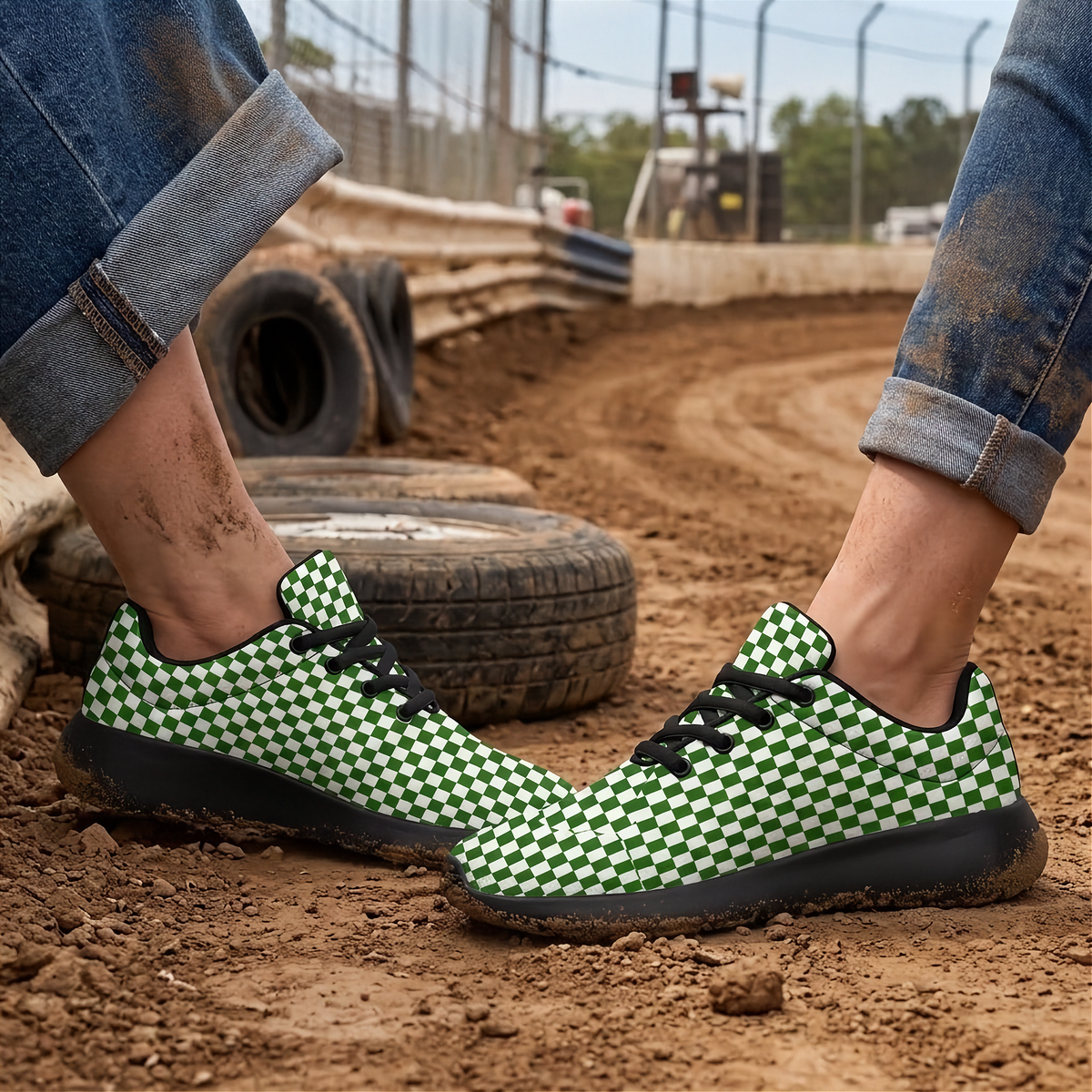 A person wearing green and black checkered sneakers on a dirt track, with cuffed blue jeans and stacked tires in the background.