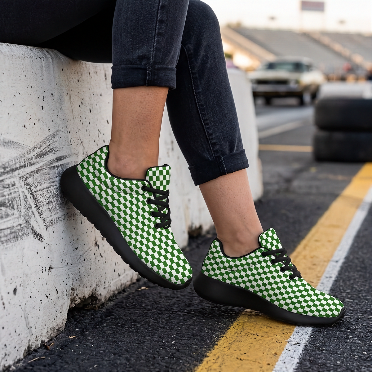 A person wearing green and black checkered low-top sneakers, sitting on a concrete barrier on an outdoor street with asphalt road and stadium seating in the background.