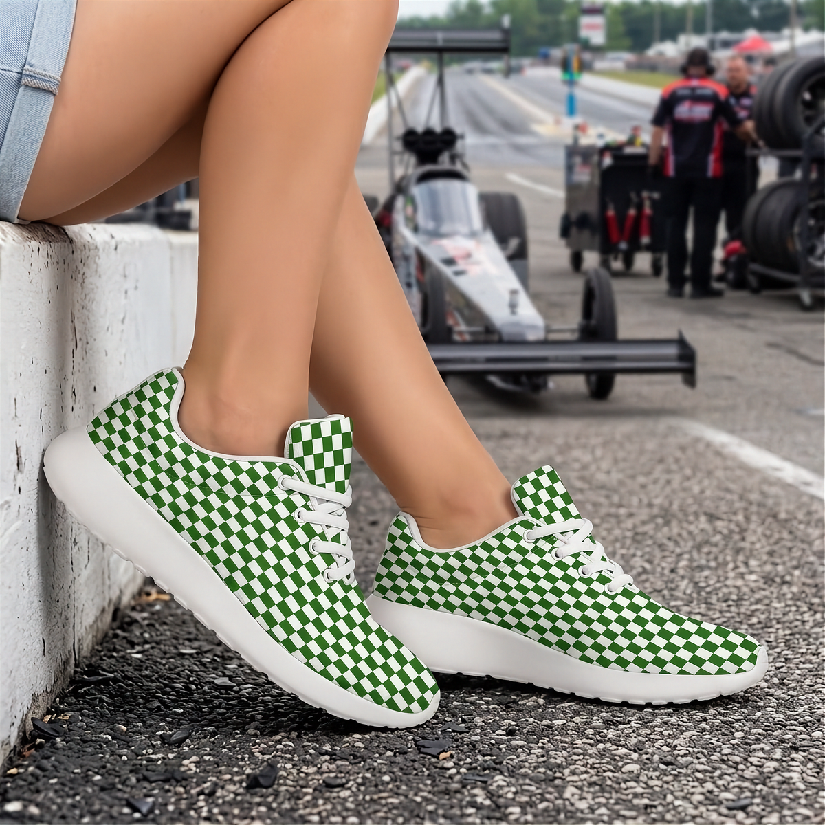 A person sitting on a white barrier at a racetrack wearing green and white checkered sneakers with white soles. A race car and track environment are blurred in the background.