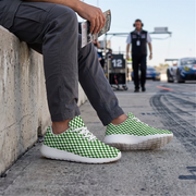 A person sitting on a concrete wall at a racetrack wearing green and white checkered sneakers with white soles. The background shows a blurred race track environment with a person standing in the distance.
