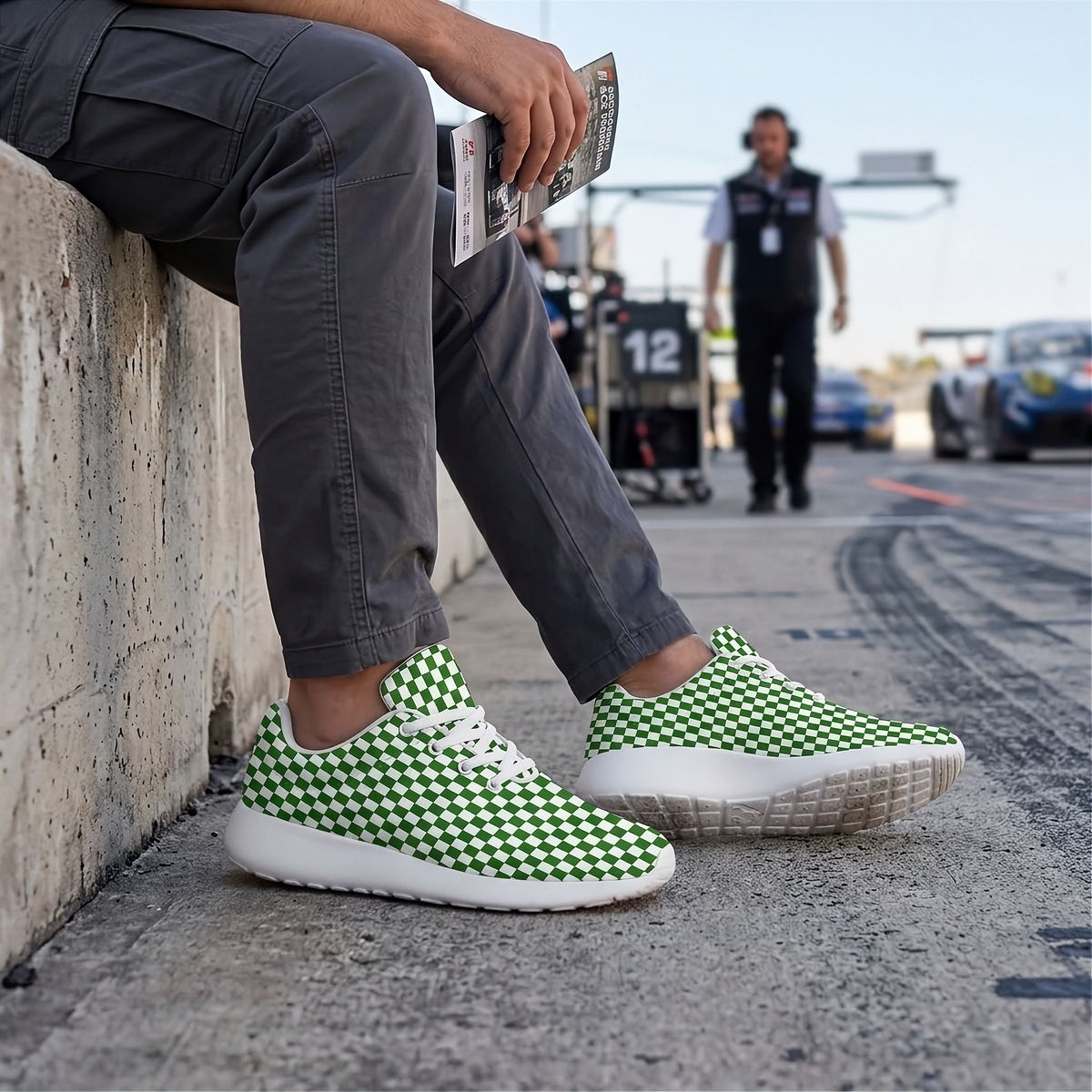 A person sitting on a concrete wall at a racetrack wearing green and white checkered sneakers with white soles. The background shows a blurred race track environment with a person standing in the distance.