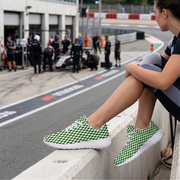 A person sitting on a concrete barrier at a racetrack wearing green and white checkered sneakers, with a blurred background showing a race car pit area and track fencing.