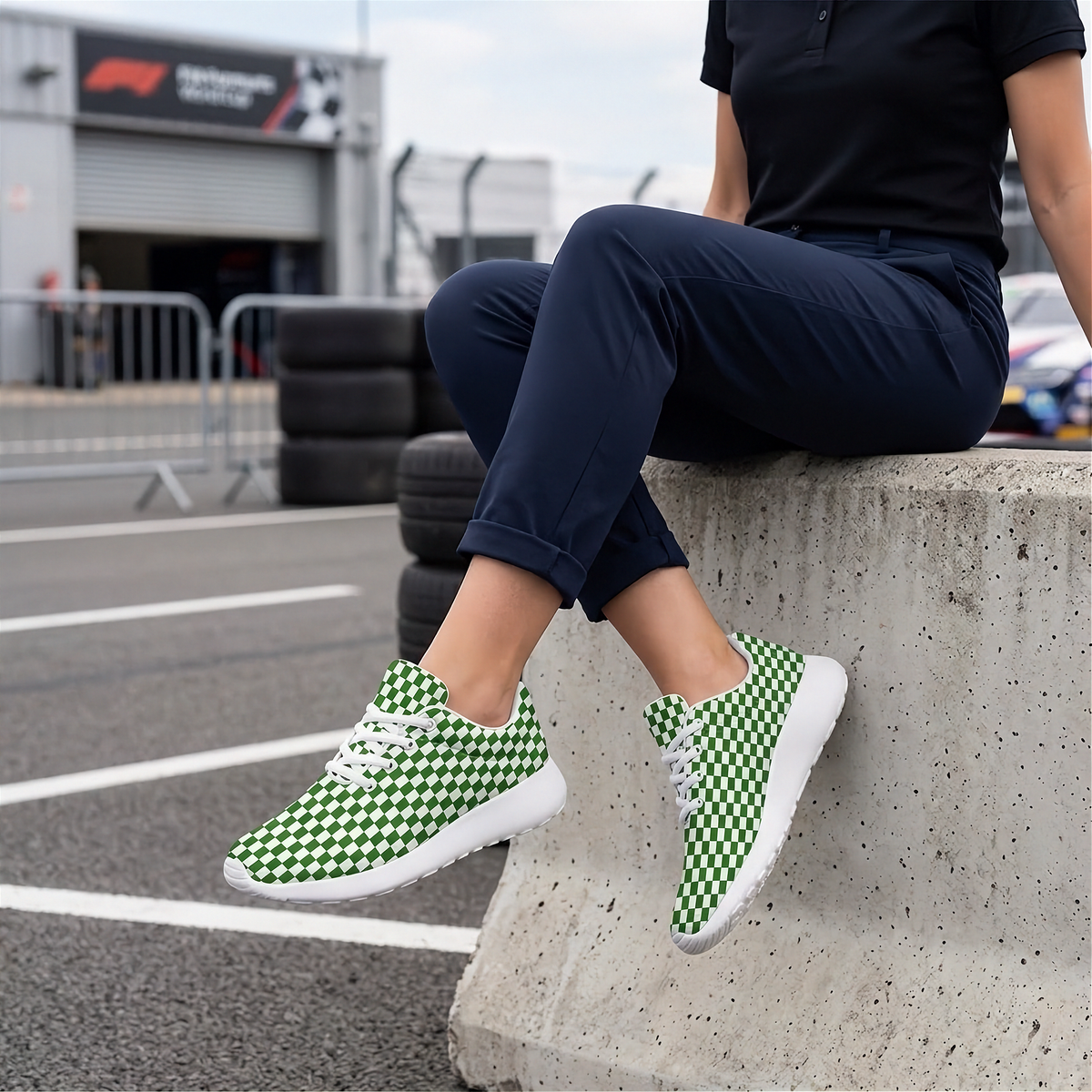 A person sitting on a concrete barrier at a racetrack wearing green and white checkered sneakers, navy pants, and a black polo shirt. The background shows a race track garage and tires.